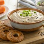 This Is How to Make the Best Creamy Bagel Dip 11 A bowl of creamy bagel dip garnished with everything bagel seasoning and green onions, served with crispy bagel chips and fresh vegetables.